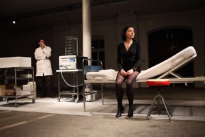 Kobieta ubrana na czarno, siedząca na łóżku szpitalnym. Obok znajduje się aparatura do transfuzji krwi oraz lekarz w kitlu, obserwujący artystkę.