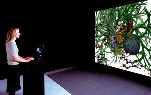 Instalacja artystyczna wykonana techniką komputerową, wyświetlana na dużym ekranie. Przed ekranem ustawiono czarny kubik z tabletem, przy którym stoi kobieta w długich włosach.